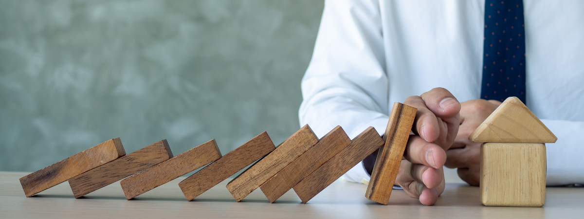 Une main protège une maison miniature d'une chute de domino représentant l'assurance en copropriété imposée par la loi ALUR