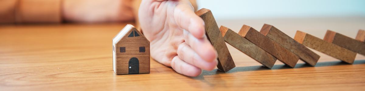 Fausse maison en bois avec une main la protégeant de dominos tombant dessus représentant la protection d'une assurance en copropriété